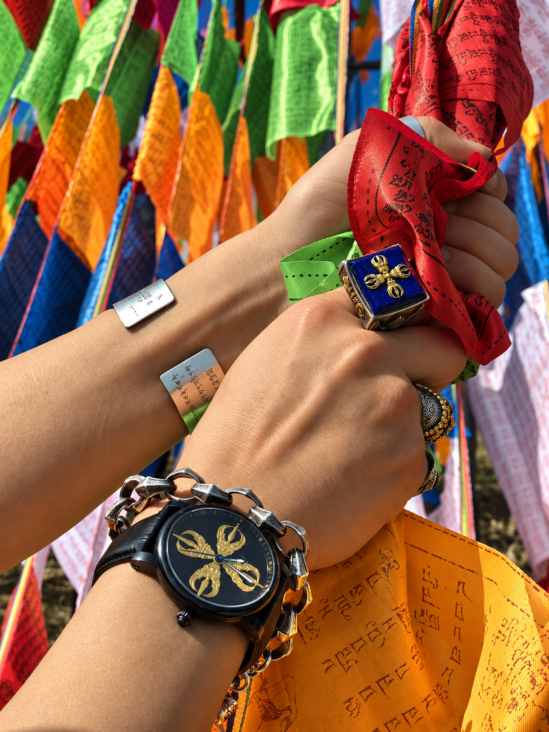 Close-up of a BuddhaHour Double Vajra watch with a black dial and golden Buddhist symbol, worn alongside traditional jewelry while holding colorful Tibetan prayer flags in natural sunlight.